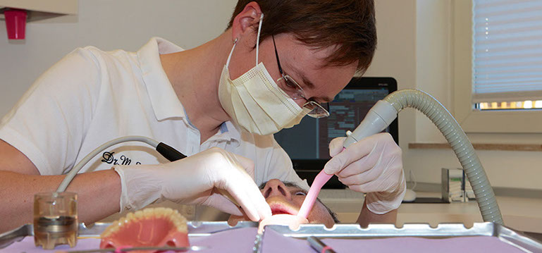 Zahnärztin behandelt Patientin im Mundbereich, auf dem Tisch der Behandlungseinheit liegen Instrumente und eine Zahnprothese